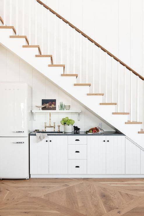 Beneath a two-toned staircase, white plank kitchenette cabinets are accented with oil rubbed bronze hardware and a black marble countertop fitted with a sink and a polished nickel deck mount faucet kit. A small white shelf is mounted to white shiplap trim over the sink and beside a white Smeg fridge.