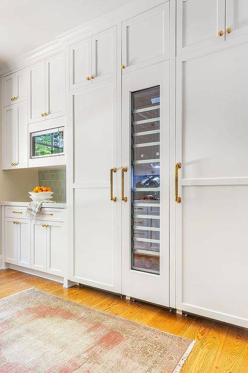 A white wood paneled refrigerator door is accented with an aged brass pull and positioned beneath white shaker cabinets and beside a custom tall glass front wine fridge.