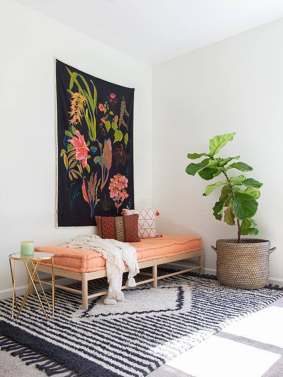 A botanical tapestry hangs over a wood daybed accented with a sherbet tufted cu،on and placed on a black ،e fringe rug between a large ،ted plant and a black and br، accent table.