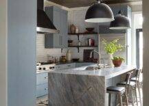 Silver and black aluminum stools sit at a gray marble waterfall edge countertop accenting a blue island finished with a sink and polished nickel faucets lit by Eugene Pendants hung from a tin ceiling,