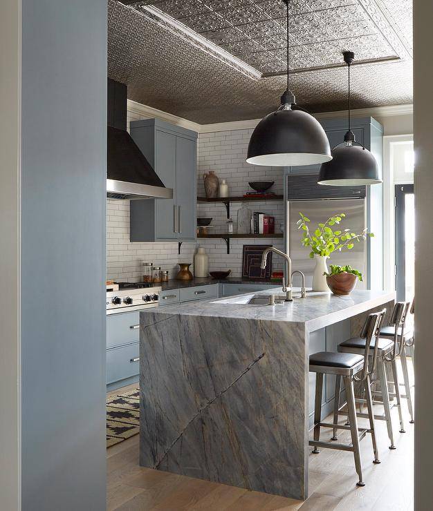 Silver and black aluminum stools sit at a gray marble waterfall edge countertop accenting a blue island finished with a sink and polished nickel faucets lit by Eugene Pendants hung from a tin ceiling,