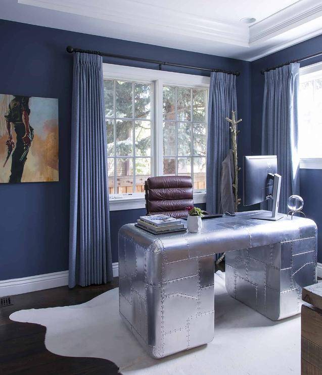 Contemporary office features an aluminum aviator desk atop a white cowhide rug, a brown leather channel tufted desk chair, blue walls with blue curtains and a silver leaf faux bois coat rack.