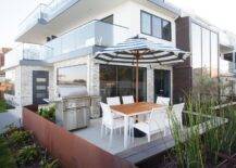 California beach style patio features a white dining table surrounded by white modern dining chairs shaded by a black and white striped umbrella while a stainless steel grill sits on a concrete floor.