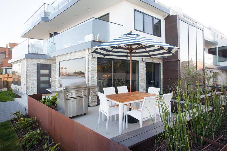 California beach style patio features a white dining table surrounded by white modern dining chairs shaded by a black and white striped umbrella while a stainless steel grill sits on a concrete floor.