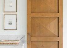 Prism blond wood door panels in a bathroom on railing mixes contemporary with a modern farmhouse appeal. Marble floors, a white washstand and brass small knobs keep the surroundings chic and neutral.