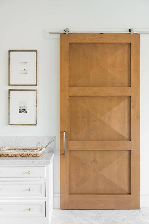 Prism blond wood door panels in a bathroom on railing mixes contemporary with a modern farmhouse appeal. Marble floors, a white washstand and brass small knobs keep the surroundings chic and neutral.