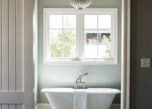 Aerin Hampton Large Pendant illuminates a cottage bathroom featuring a white freestanding rolled top bathtub under a window. Gray plank barn doors mounted on black metal railing adds to a cottage theme creating a soothing and restful bathroom design.