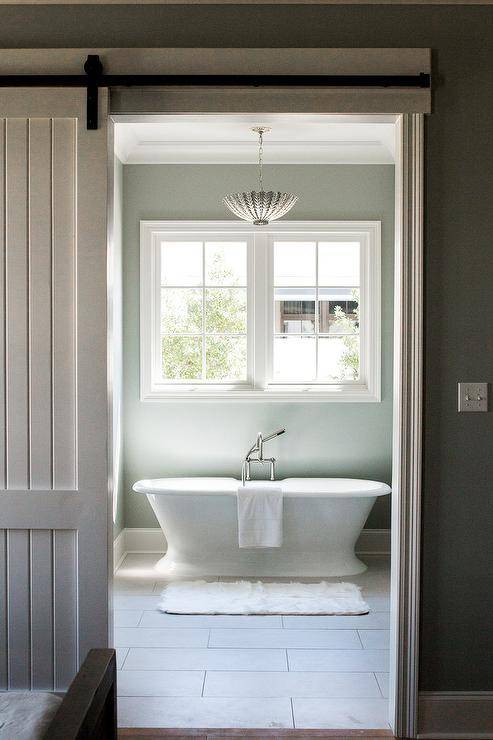 Aerin Hampton Large Pendant illuminates a cottage bathroom featuring a white freestanding rolled top bathtub under a window. Gray plank barn doors mounted on black metal railing adds to a cottage theme creating a soothing and restful bathroom design.