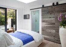 Gorgeous bedroom with white slipcovered full sized bed layered with crisp white linens and a royal blue coverlet alongside iron french doors leading to a courtyard patio. The bedroom features rustic sliding barn doors which open to reveal a glass front shower with white tile surround and brass shower head.