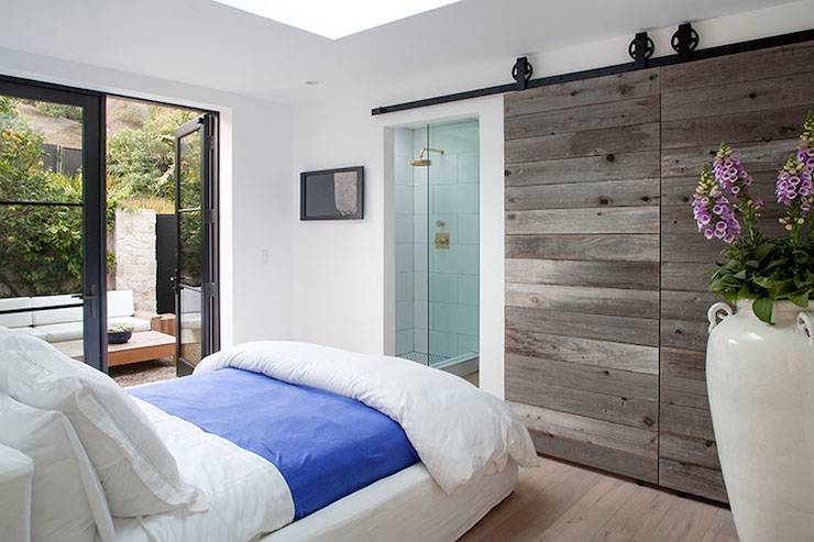 Gorgeous bedroom with white slipcovered full sized bed layered with crisp white linens and a royal blue coverlet alongside iron french doors leading to a courtyard patio. The bedroom features rustic sliding barn doors which open to reveal a glass front shower with white tile surround and brass shower head.