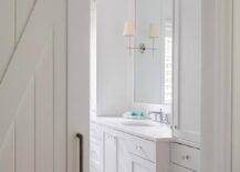 White shiplap barn doors slide open into a master bathroom showcasing a vanity mirror flanked by Thomas O'Brien Bryant Sconces. Black and white vintage hex floor tiles add a contrast to white shaker cabinets, white quartz countertops, and glass knobs.