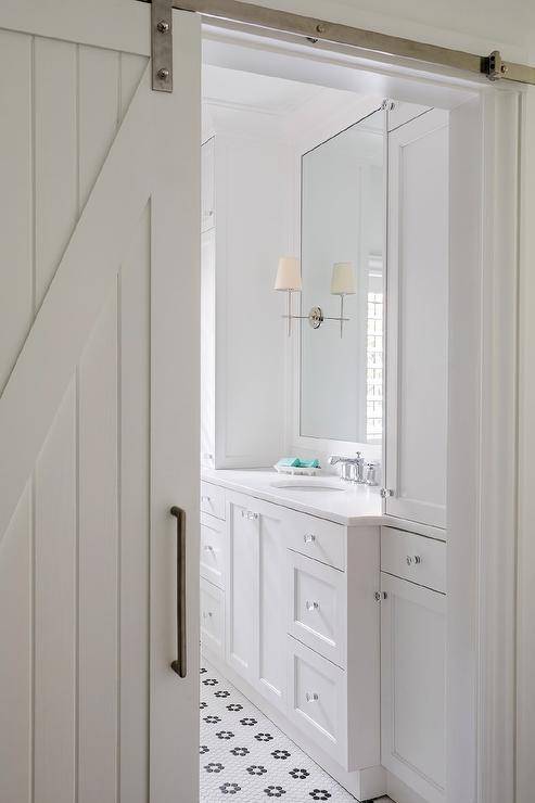 White shiplap barn doors slide open into a master bathroom showcasing a vanity mirror flanked by Thomas O'Brien Bryant Sconces. Black and white vintage hex floor tiles add a contrast to white shaker cabinets, white quartz countertops, and glass knobs.