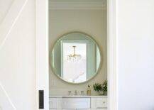 A white barn door on rails opens to an en suite bathroom boastin ga white curved bath vanity fixed beneath a round gold mirror.