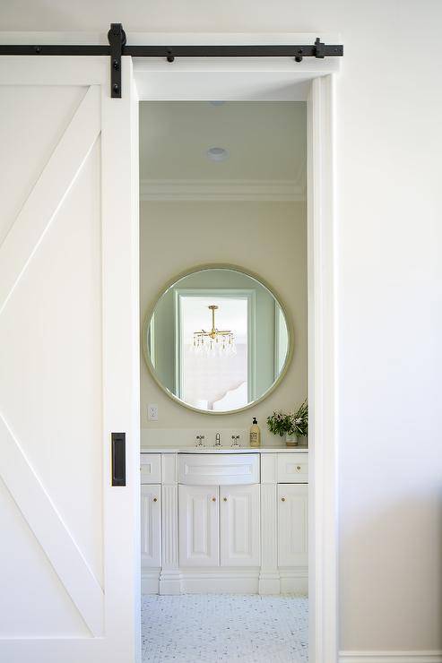 A white barn door on rails opens to an en suite bathroom boastin ga white curved bath vanity fixed beneath a round gold mirror.