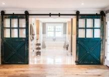 Fabulous master bathroom with wide planked hardwood floors alongside shiplap clad walls framing doorway highlighted by pair of peacock blue sliding barn doors with glass panes. The bathroom features glossy white tiled floors alongside subway tiled lower walls with taupe paint color on upper walls accentuating a double sash window over the pedestal tub with floor mount faucet.