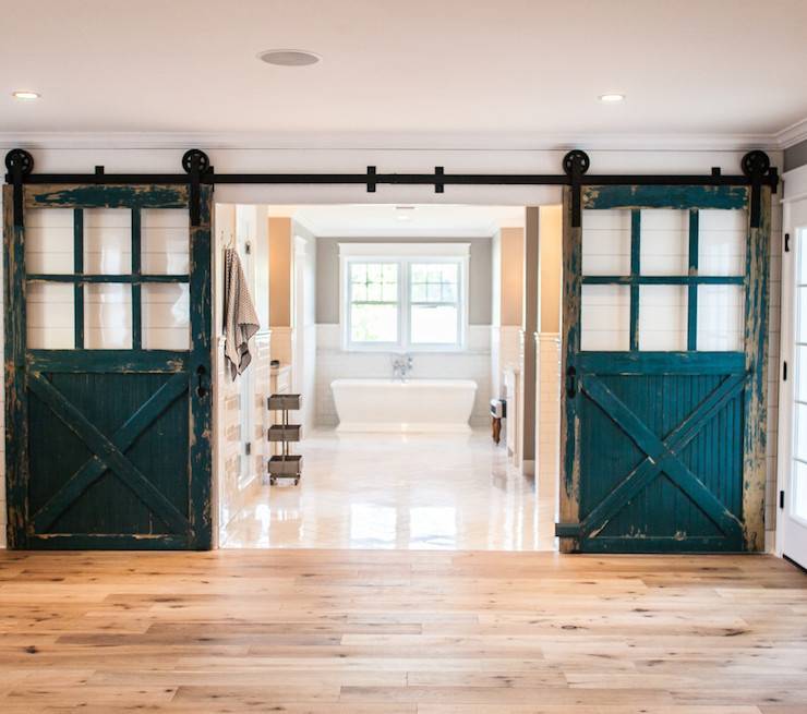 Fabulous master bathroom with wide planked hardwood floors alongside shiplap clad walls framing doorway highlighted by pair of peacock blue sliding barn doors with glass panes. The bathroom features glossy white tiled floors alongside subway tiled lower walls with taupe paint color on upper walls accentuating a double sash window over the pedestal tub with floor mount faucet.