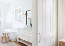 A white barn door on rails slides open to reveal a master bathroom boasting a pink and brown vintage rug placed in front of a white dual bath vanity located beside an oval bathtub. The bathtub is matched with a rustic stool and located beneath a window.