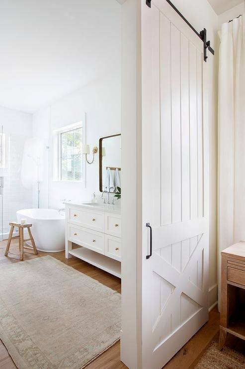 A white barn door on rails slides open to reveal a master bathroom boasting a pink and brown vintage rug placed in front of a white dual bath vanity located beside an oval bathtub. The bathtub is matched with a rustic stool and located beneath a window.