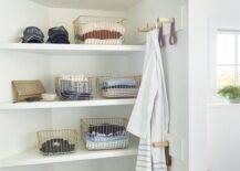 White bathroom shelves are stacked in a corner over gold wire baskets and adjacent to leather and wooden peg towel hooks.