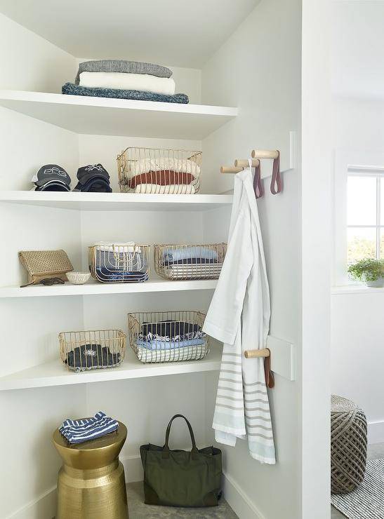 White bathroom shelves are stacked in a corner over gold wire baskets and adjacent to leather and wooden peg towel hooks.