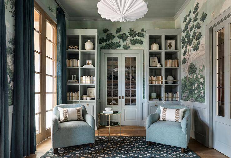 Placed on a blue lattice rug, blue boucle chairs complemented with a round glass and hammered brass accent table sit in front of blue built-in bookshelves with brass trim. The bookshelves are mounted against a wall clad in French hand painted wallpaper and flank blue double doors with glass panels.