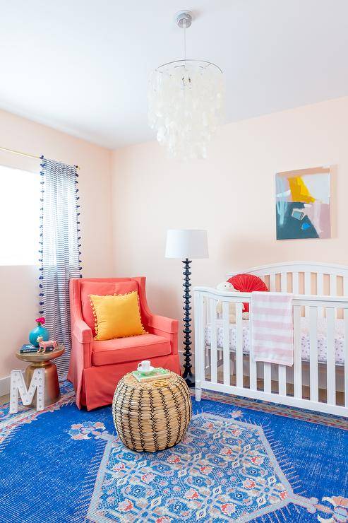 Orange and blue nursery boasts a bright blue vintage rug with a woven round pouf and a hot pink accent chair beside a white wooden crib. A white capiz chandelier adds a contemporary finish to the sweet nursery space.