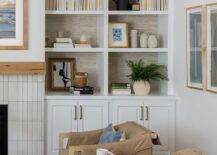 Brown leather flap accent chairs topped with blue pillows sit on a blue stripe jute rug on either side of a round wooden accent table. Gray wallpaper accents the back of an arched built-in bookcase mounted over white cabinets.