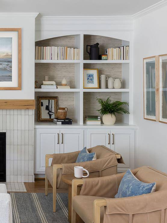 Brown leather flap accent chairs topped with blue pillows sit on a blue stripe jute rug on either side of a round wooden accent table. Gray wallpaper accents the back of an arched built-in bookcase mounted over white cabinets.