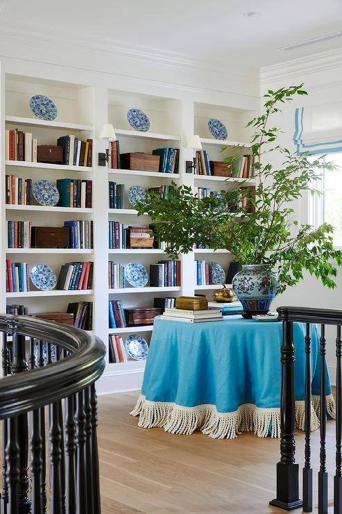 Staircase landing library features an azure blue fringe skirted table with potted plant and built in bookshelves.