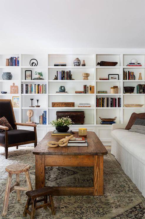 A wall of built-in bookshelves is fixed to the side of a white slipcovered sofa placed on a gray and green rug at a reclaimed wood coffee table complemented with rustic wooden stools.