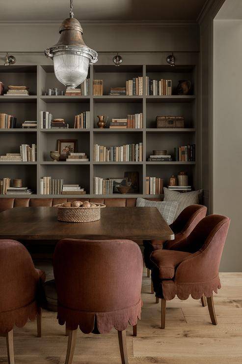 Red scalloped slipcovered dining chairs sit around a brown wooden dining table lit by a vintage lantern hung from a gray painted ceiling and placed on a wide plank wood floor in front of gray bookshelves built into a gray wall.