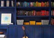 Home office features bold blue bookshelves with books organized by color lit by bronze picture lights and paint splatter accent pillows.
