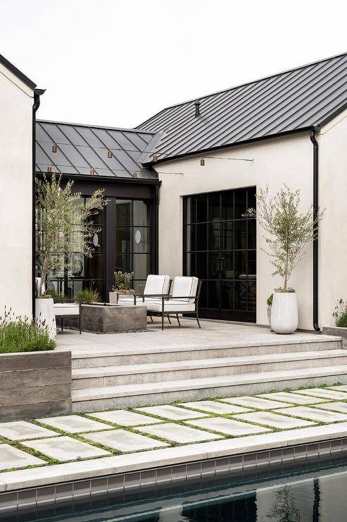 Concrete pavers are fixed over grass in front of steps leading to a plank fire pit flanked by black aluminum lounge chairs contrasted with white cushions.