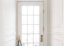 A glass paneled white door with a brass door knob and plate is located in a vestibule beneath an arched transom window. A brass Hicks pendant is hung between white walls with white trim and over a dark stained wood floor.