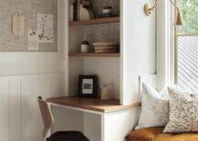 Brown wooden shelves are mounted over a wood top kids desk matched with a wood desk chair and fixed beside a storage window seat lit by brass swing arm sconces fixed above an orange tufted cushion.