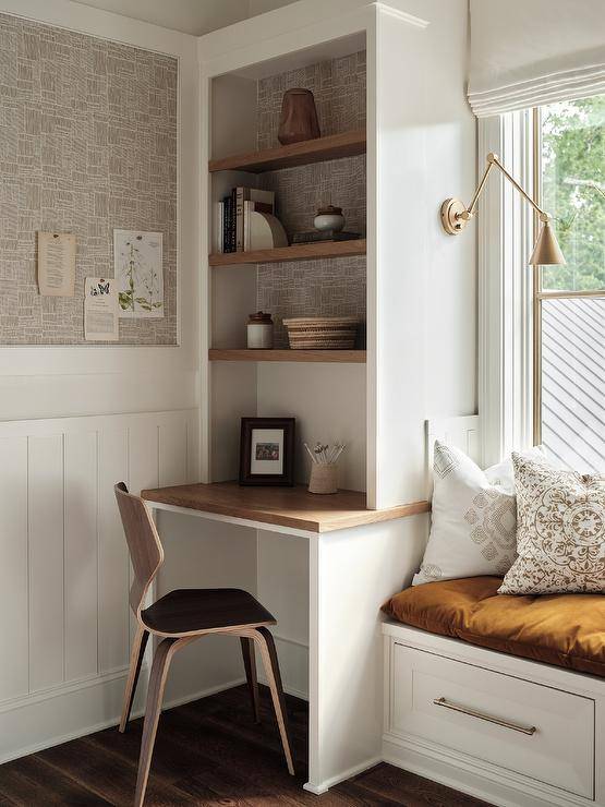 Brown wooden shelves are mounted over a wood top kids desk matched with a wood desk chair and fixed beside a storage window seat lit by brass swing arm sconces fixed above an orange tufted cushion.