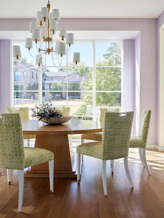 A glass and brass chandelier hangs over a round brown pedestal dining table paired with white and green damask dining chairs, while purple wallpaper frames a large window.