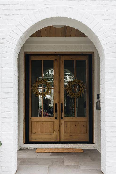 A white painted brick home features wood and glass double front doors accented with wreaths and oil rubbed bronze door handles.