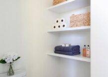 A minimalist bathroom boasts a nook filled with stacked white floating shelves lined with wicker baskets and folded towels.