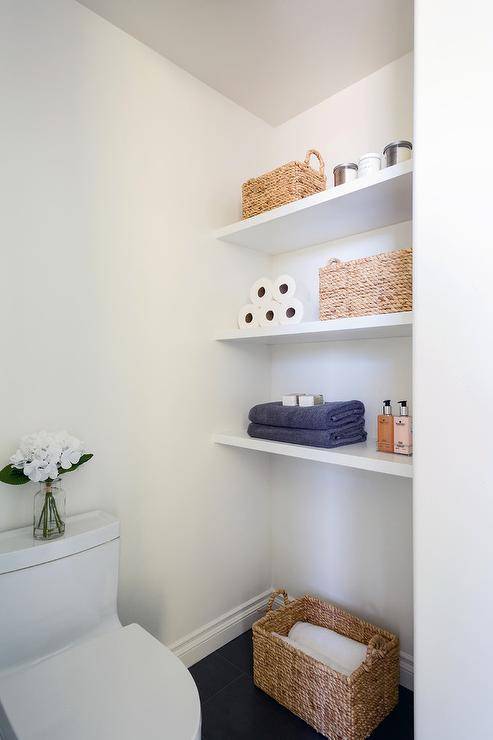 A minimalist bathroom boasts a nook filled with stacked white floating shelves lined with wicker baskets and folded towels.