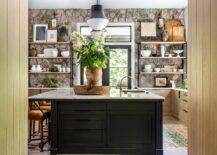 A stunning rustic stone wall accent an eclectic kitchen boasting wood and metal shelves fixed on either side of a black French doors located beneath a transom window. Burnt orange velvet stools sit at a dark brown wood island finished with a sink and lit by two Kelly Wearstler Precision pendants.