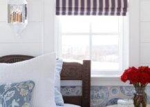 Beautiful French country bedroom boasts a carved wooden bed dressed in white and blue bedding topped with whtie and blue pillows. The bed is positioned against a wal clad in blue French plate print wallpaper flanking a white plank trim. A window is covered in a red, white, and blue striped roman shade.