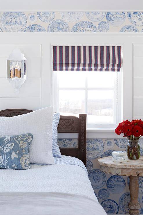 Beautiful French country bedroom boasts a carved wooden bed dressed in white and blue bedding topped with whtie and blue pillows. The bed is positioned against a wal clad in blue French plate print wallpaper flanking a white plank trim. A window is covered in a red, white, and blue striped roman shade.