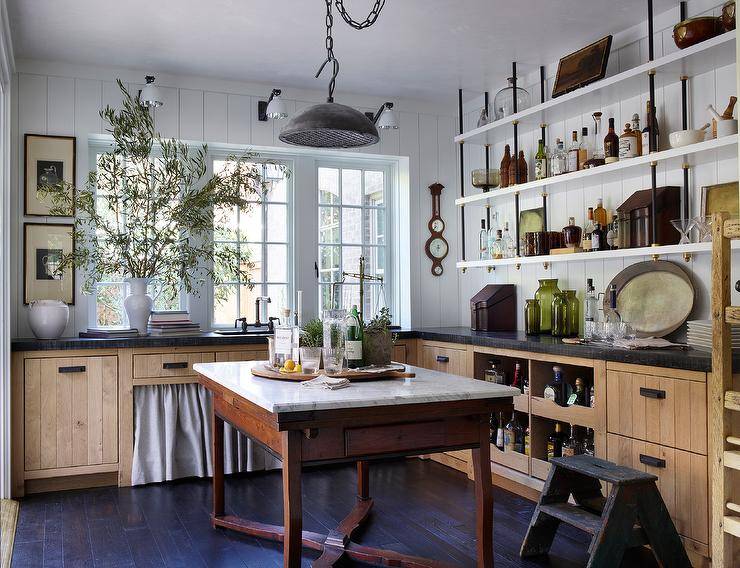 French country kitchen features an antique French island fitted with a marble top and placed on an ebony stained wood floor. French shelves are stacked in front of white plank trim and over rustic cabinets finished with pull-out shelves, oil rubbed bronze hardware, and a black leathered countertop. A row of windows is positioned over a sink located above skirted cabinets.