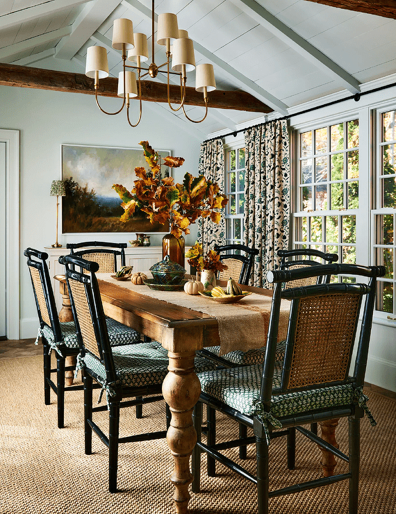 Dining room features black bamboo and cane dining chairs at a French dining table with turned legs lit by a brass Vendome chandelier hung from a vaulted plank ceiling and a tan rug.