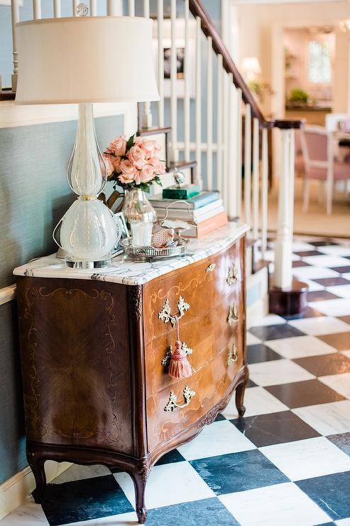 Black and white marble harlequin floor tiles bring a charming old-world finish to a foyer designed with a French dresser with pink tassels displaying a light blue glass lamp. The French dresser boasts styled decor against a staircase wall finished with green grasscloth wallpaper.
