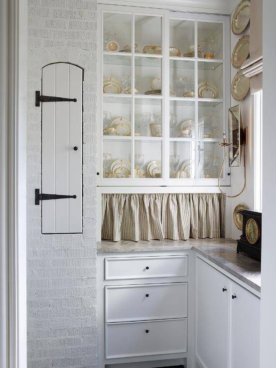 Wonderfully appointed French country kitchen pantry features glass cabinets finished with a gray skirt and positioned above white cabinets donning small oil rubbed bronze knobs.