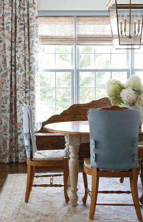 French country dining room features rush seat dining chairs with blue backs placed on a cream vintage rug at a French country oval dining table. The table is lit by a brass lantern, as floral curtains hang in front of bamboo roman shades.