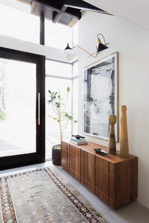 Gold and black sconces illuminate a large black and white abstract art piece hung above a brown mid-century modern credenza. A brown and blue vintage runner leads to a black front door finished with one large glass panel and flanked by sidelights.