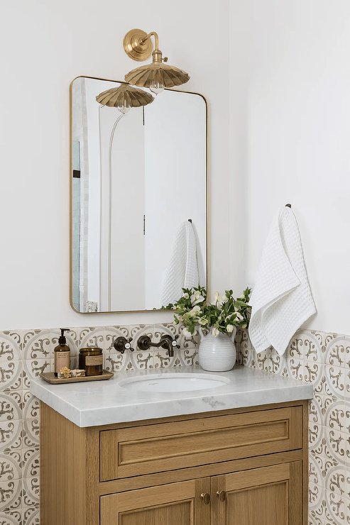 A powder room wall half clad in white and brown mosaic tiles features a brown single washstand donning brass knobs and a marble countertop holding a sink beneath a wall mount oil rubbed bronze vintage faucet. The faucet is fixed beneath a curved brass mirror illuminated by a vintage brass sconce.